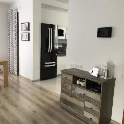 Jasne panele podłogowe w salonie z widokiem na kuchnię, czarna lodówka i komoda w stylu rustykalnym.