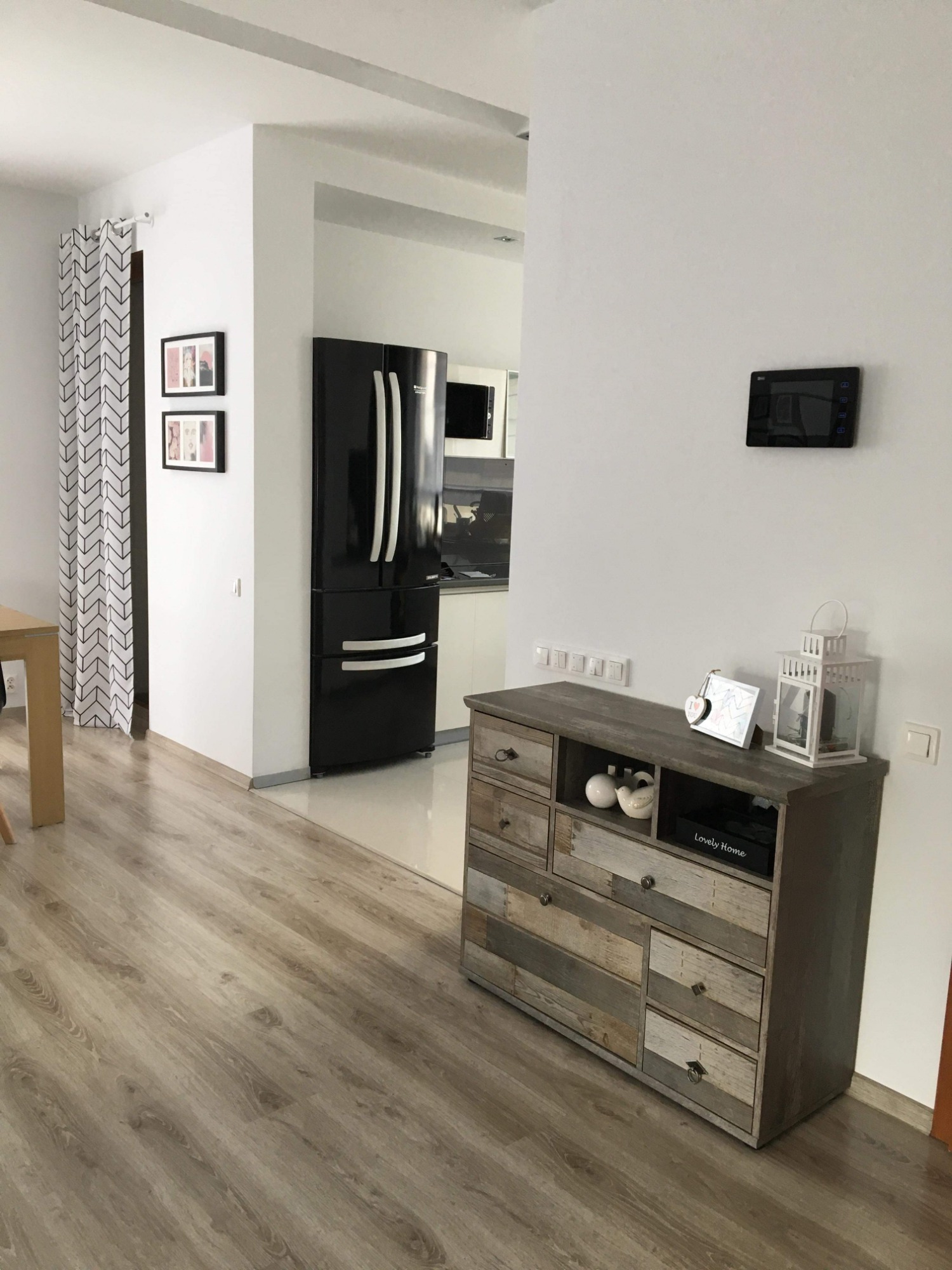 Jasne panele podłogowe w salonie z widokiem na kuchnię, czarna lodówka i komoda w stylu rustykalnym.