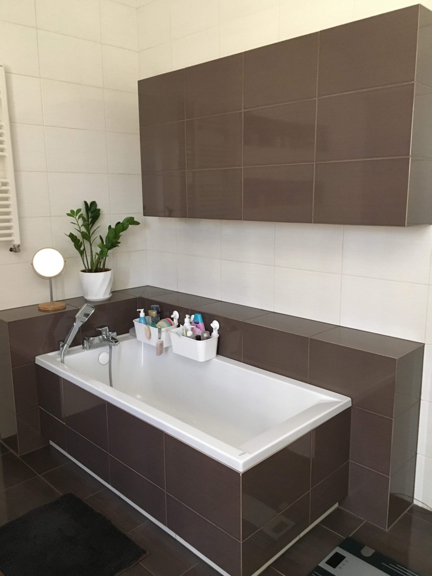 Wykończona łazienka z brązowo-białymi płytkami, białą wanną obudowaną brązowymi płytkami, rośliną w doniczce, lusterkiem i organizerami z kosmetykami.