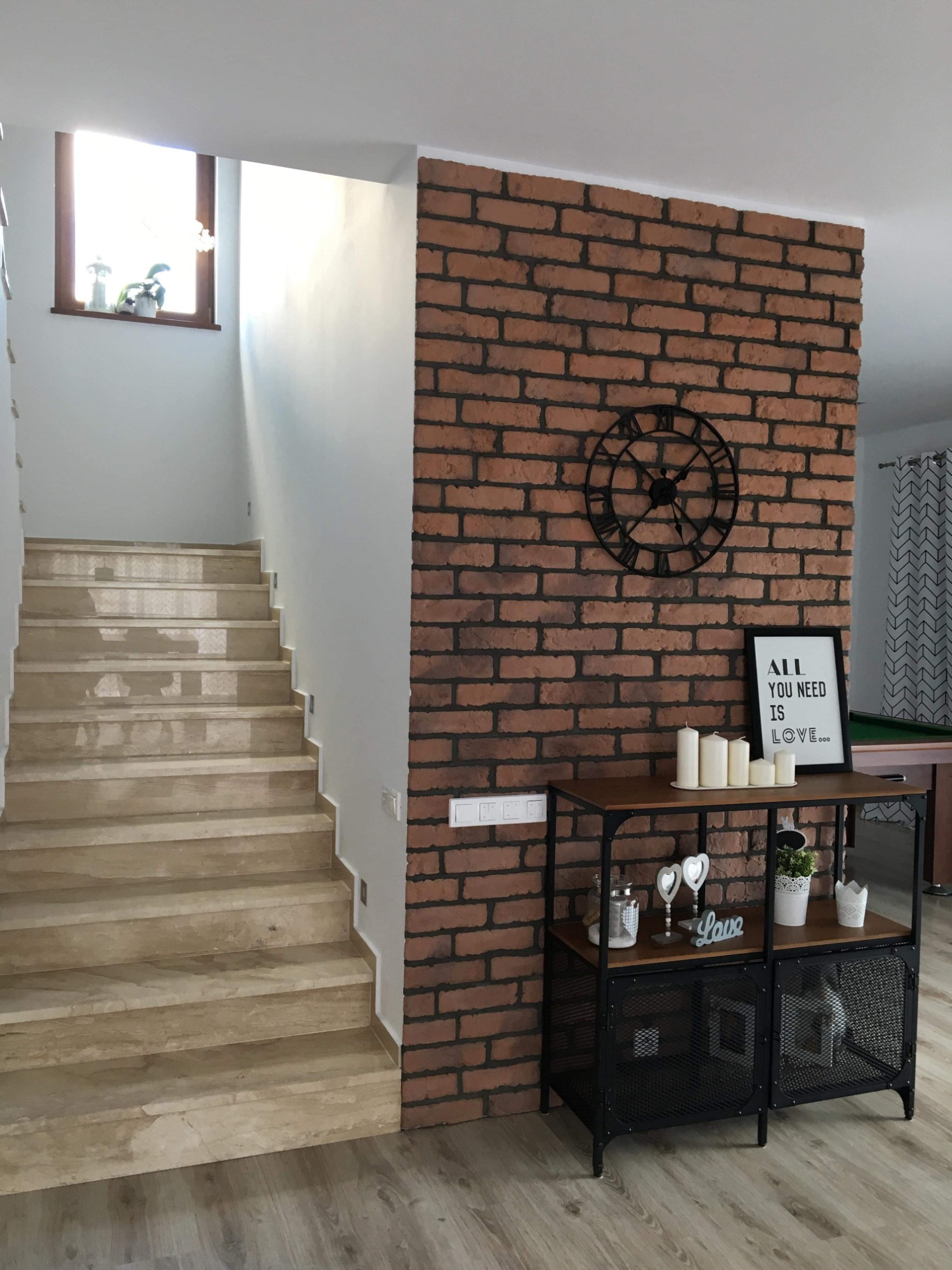 Klatka schodowa z marmurowymi stopniami, ściana z cegły klinkierowej z dekoracyjnym zegarem i konsolą z dekoracjami, widok na salon z bilardem w tle.