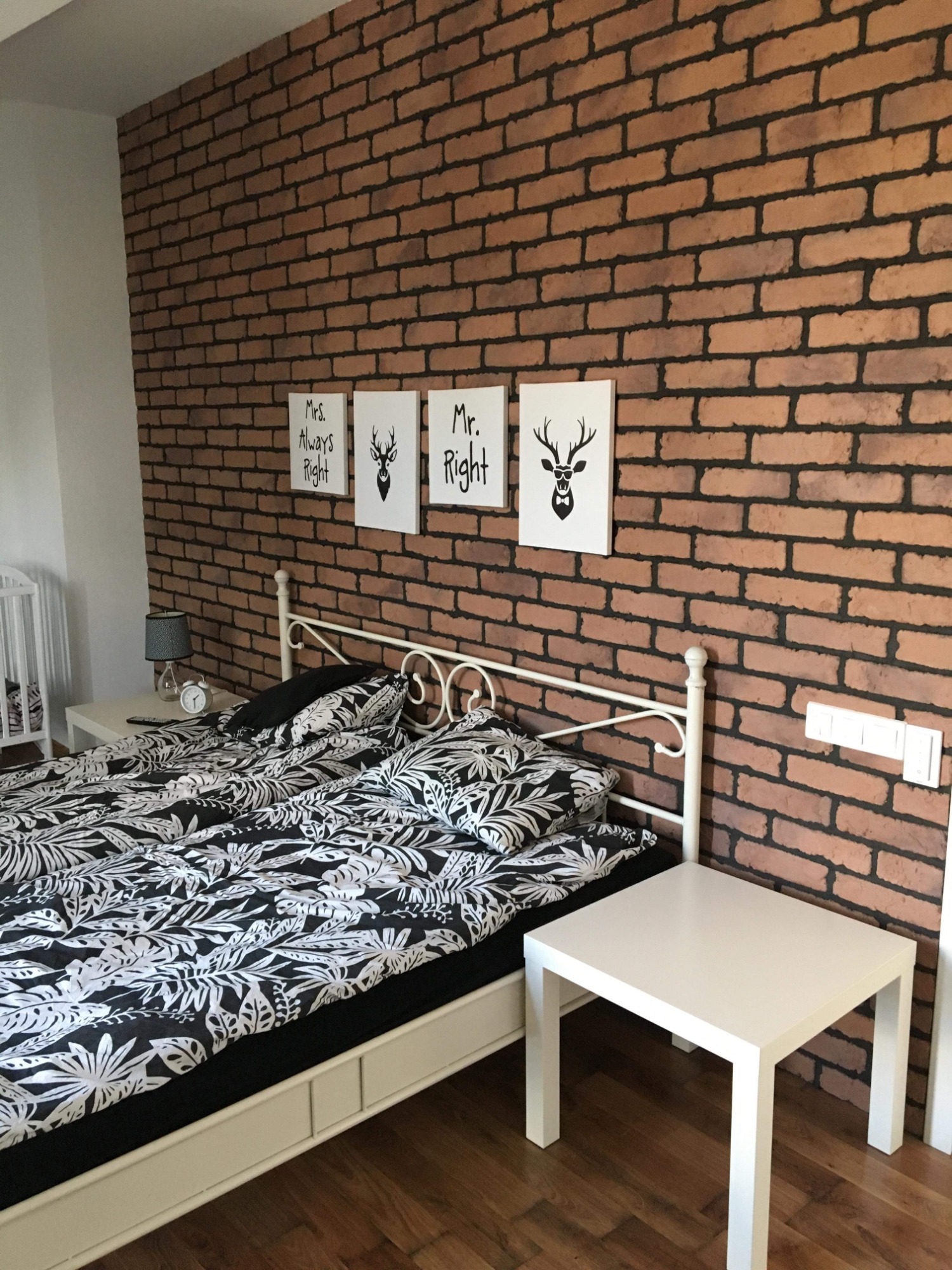 Sypialnia z dekoracyjną ścianą z cegły, białym łóżkiem z pościelą w liście, białymi stolikami nocnymi i obrazkami na ścianie.