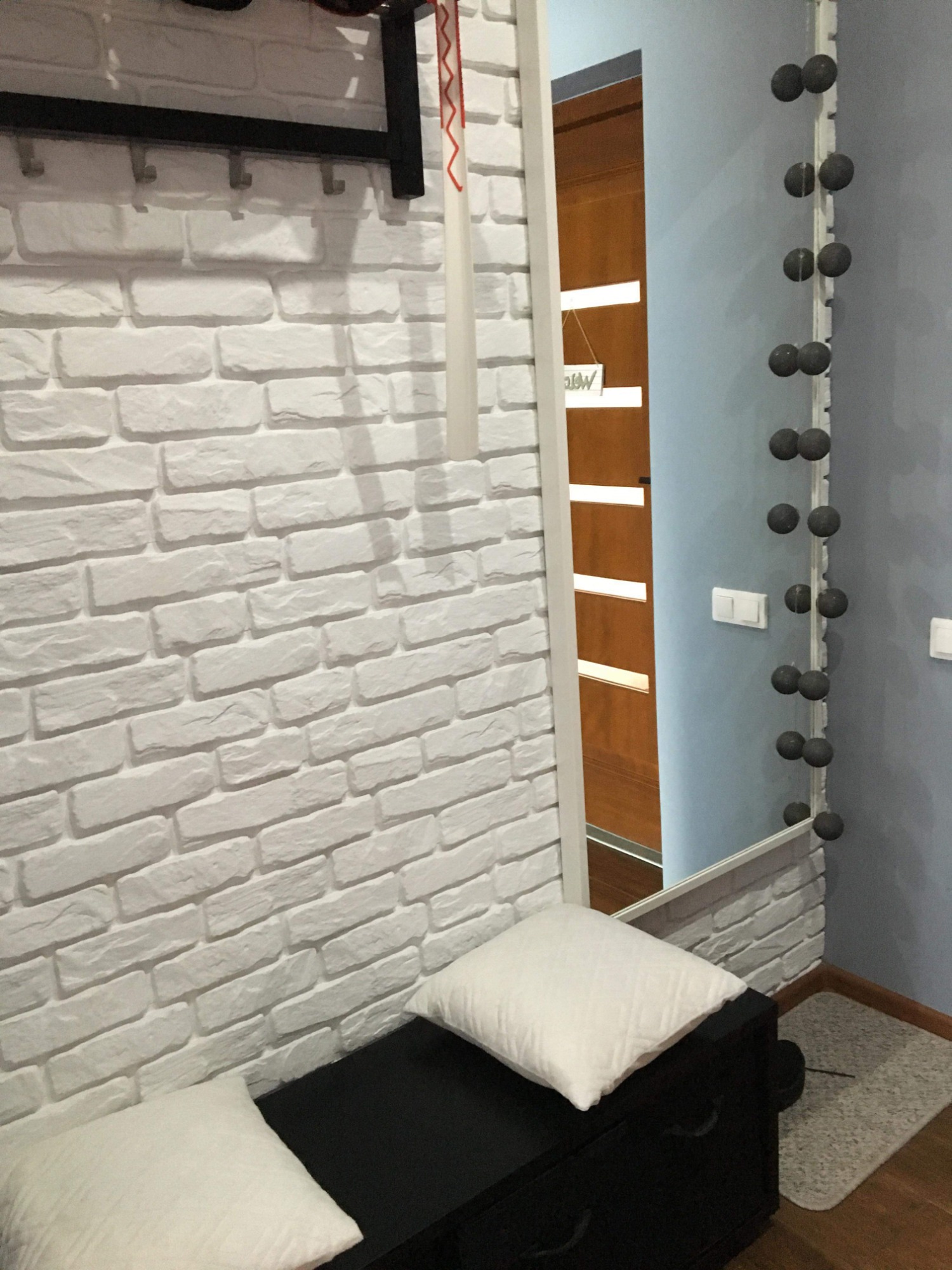 Fragment przedpokoju z białą ścianą imitującą cegłę, czarnym wieszakiem z haczykami, lustrem odbijającym drzwi i niebieską ścianę, oraz czarną ławeczką z białymi poduszkami.