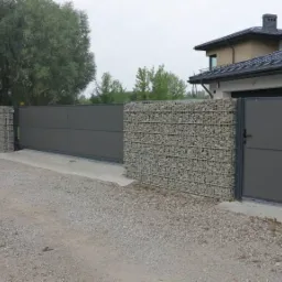 Nowoczesne ogrodzenie posesji z przesuwną bramą i furtką, z elementami gabionowymi wypełnionymi kamieniami, na tle domu z ciemną dach&oacute;wką.