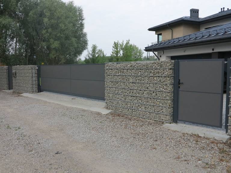 Nowoczesne ogrodzenie posesji z przesuwną bramą i furtką, z elementami gabionowymi wypełnionymi kamieniami, na tle domu z ciemną dachówką.
