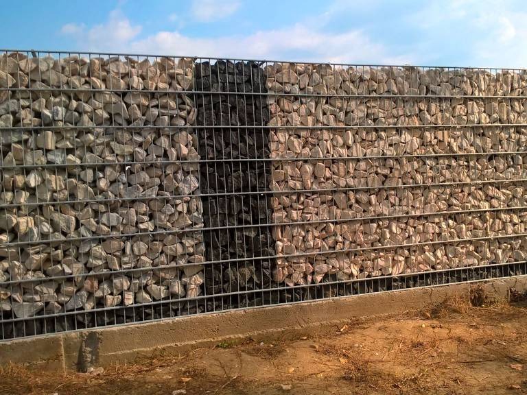 Gabionowe ogrodzenie z szarego kamienia, fragment wypełniony czarnym kamieniem, widoczne stalowe kosze i betonowa podstawa.