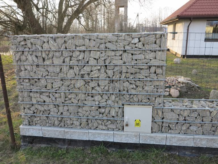 Gabionowa konstrukcja ogrodzeniowa wypełniona kamieniami, osadzona na betonowej podstawie, z widoczną skrzynką elektryczną z symbolem ostrzegawczym na froncie, w tle dom jednorodzinny i drzewa.