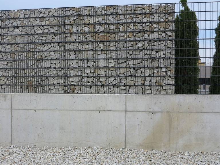 Ogrodzenie z gabionów wypełnionych kamieniami, z betonową podmurówką i fragmentem siatki ogrodzeniowej w tle, widoczne tuje.