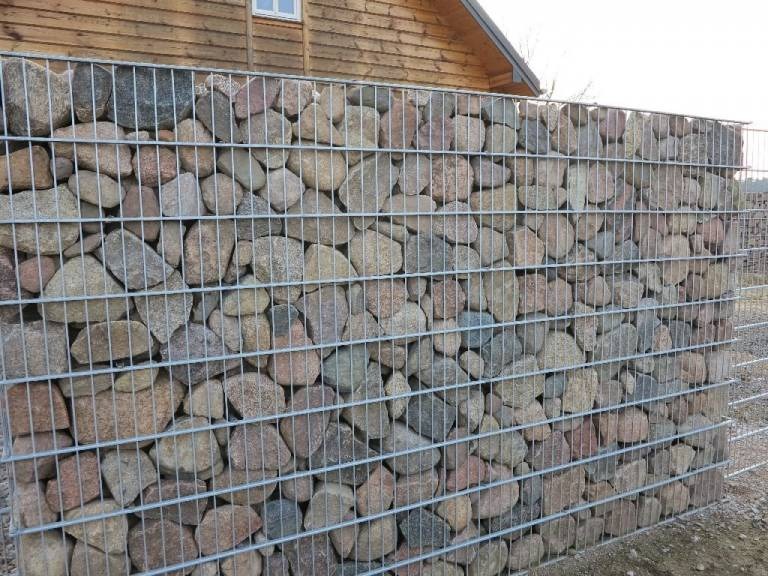 Ogrodzenie gabionowe z kamieni rzecznych w metalowej siatce na tle drewnianego domu z białym oknem.