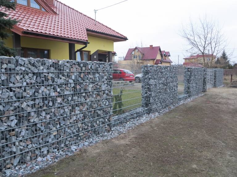 Gabionowe ogrodzenie z siatki wypełnione szarymi kamieniami, oddzielające posesję z żółtym domem i czerwonym dachem od drogi.