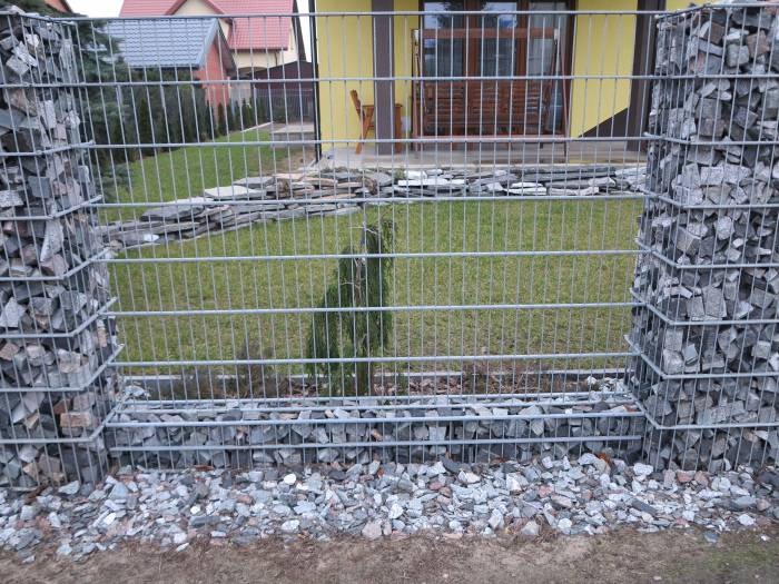 Ogrodzenie gabionowe z szarej siatki i kamieni, widok na trawnik i żółty dom w tle.