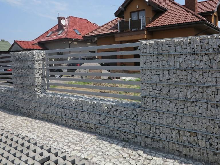 Ogrodzenie z gabionów i metalowych przęseł na tle domu z czerwoną dachówką i brukowanym podjazdem.