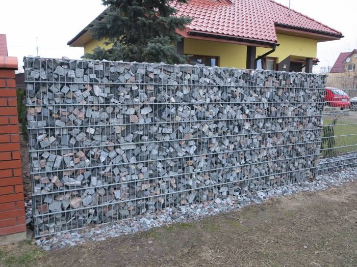 Ogrodzenie z gabionów wypełnione szarym kamieniem, z widocznym fragmentem ceglanego słupka i żółtego domu w tle.