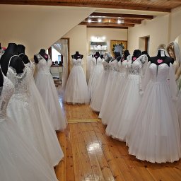 ESTER-MODA ŚLUBNA - Salon sukien ślubnych: Rząd białych sukien na manekinach, drewniana podłoga, subtelne oświetlenie, różowe serduszka z cenami na sukniach, manekin z rudymi włosami.