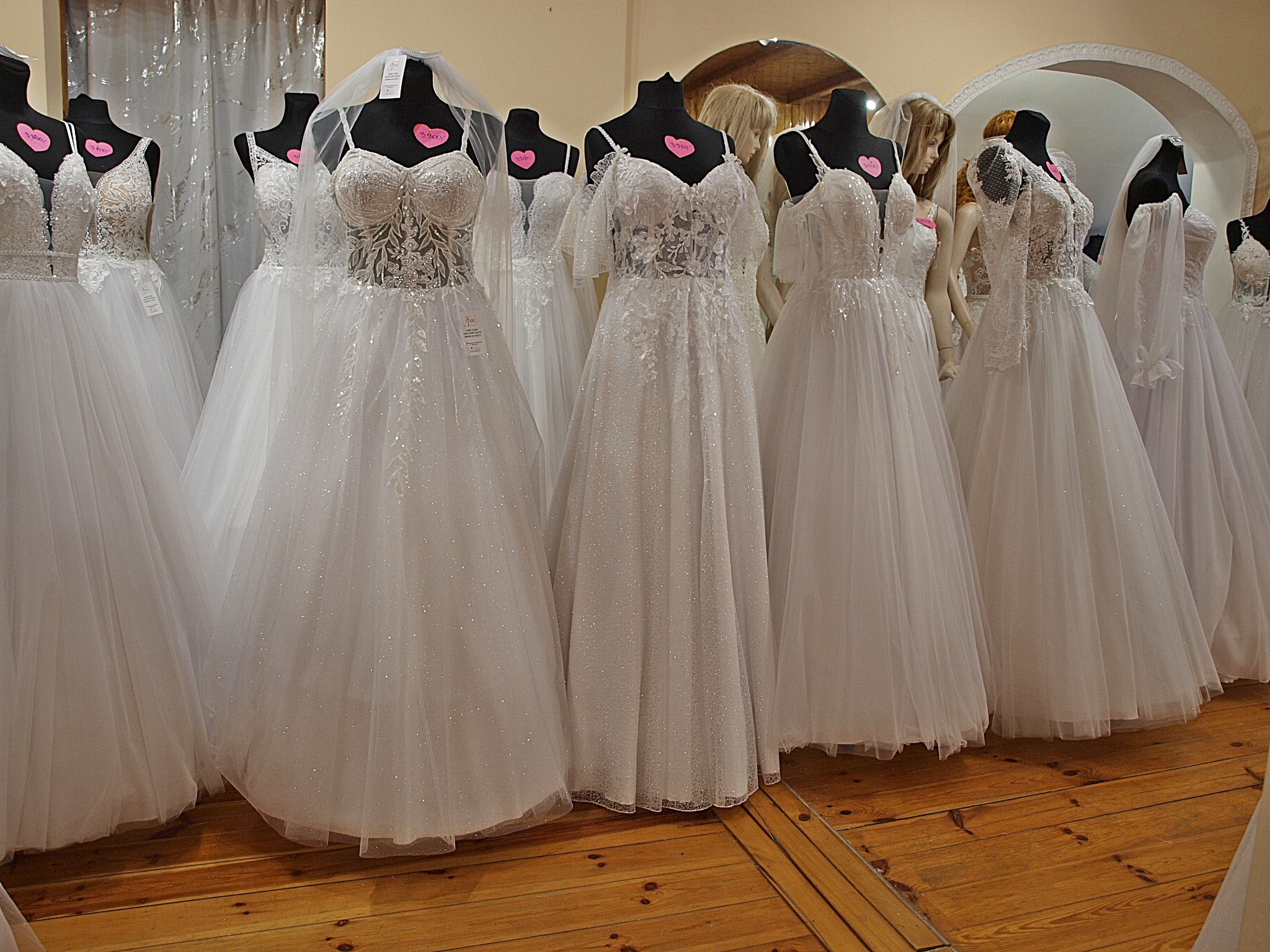 Salon sukien ślubnych: manekiny w białych, zdobionych sukniach z tiulu, ustawione na drewnianej podłodze. W tle lustro i zasłony.
