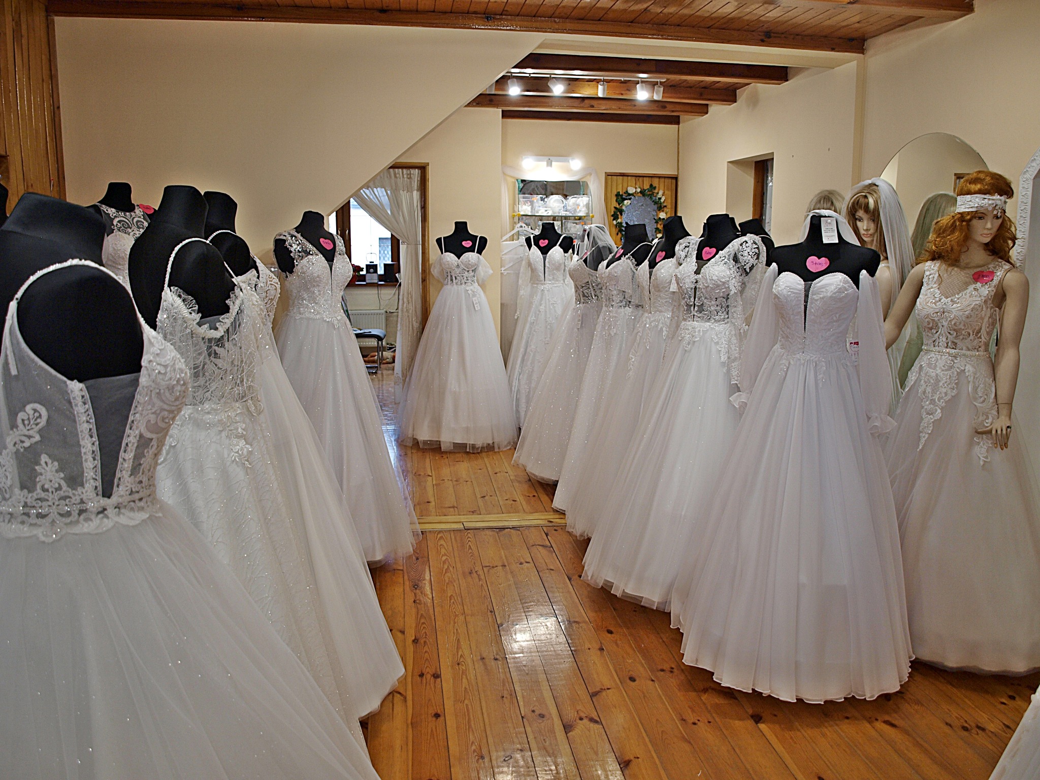 Salon sukien ślubnych: Rząd białych sukien na manekinach, drewniana podłoga, subtelne oświetlenie, różowe serduszka z cenami na sukniach, manekin z rudymi włosami.
