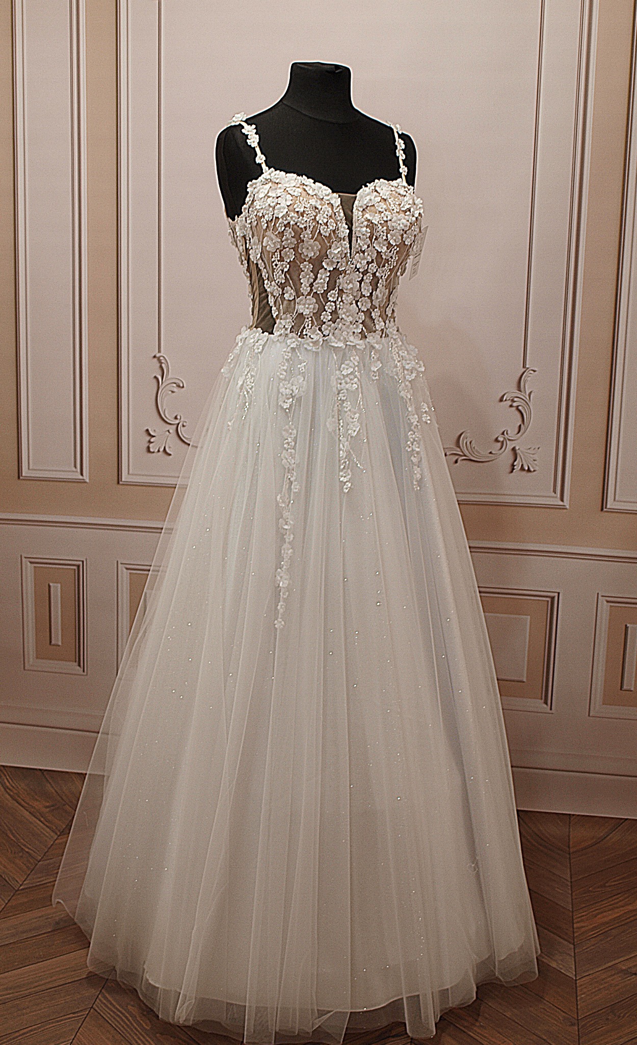 Elegancka suknia ślubna z koronkową górą, umieszczona na manekinie na tle dekoracyjnej ściany i drewnianej podłogi. Delikatne zdobienia i subtelny blask tiulu.