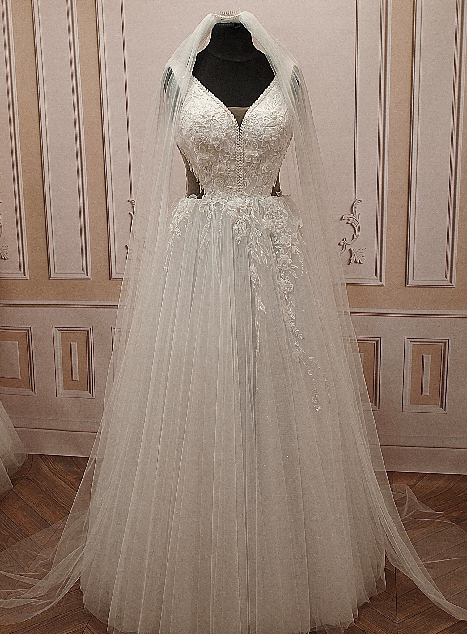 Elegancka, biała suknia ślubna z koronkową górą i tiulowym dołem, prezentowana na manekinie z welonem, na tle ściany w stylu klasycznym.