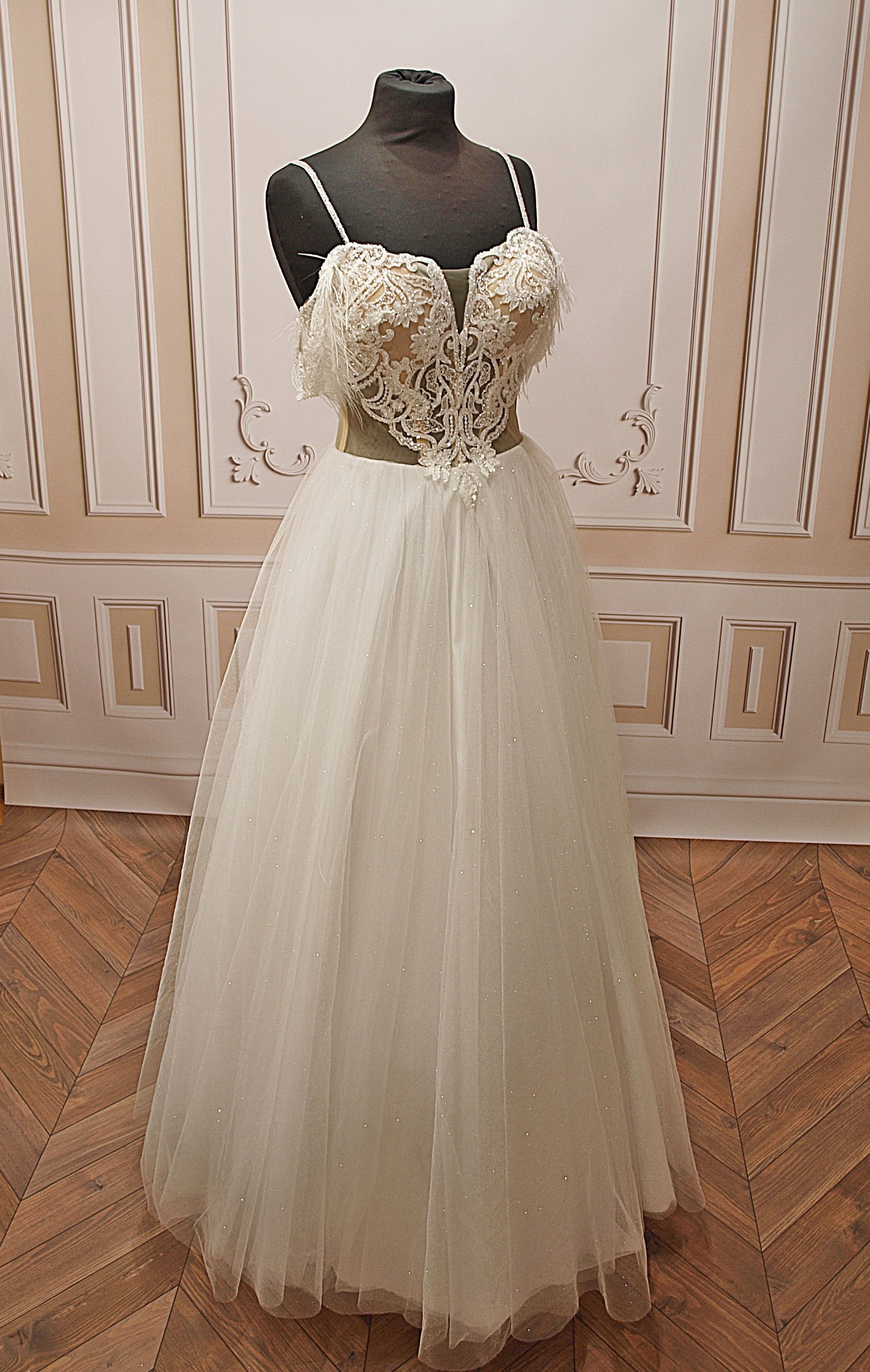 Elegancka suknia ślubna z koronkowym gorsetem i tiulowym dołem, prezentowana na manekinie na tle dekoracyjnej ściany i parkietu w jodełkę.
