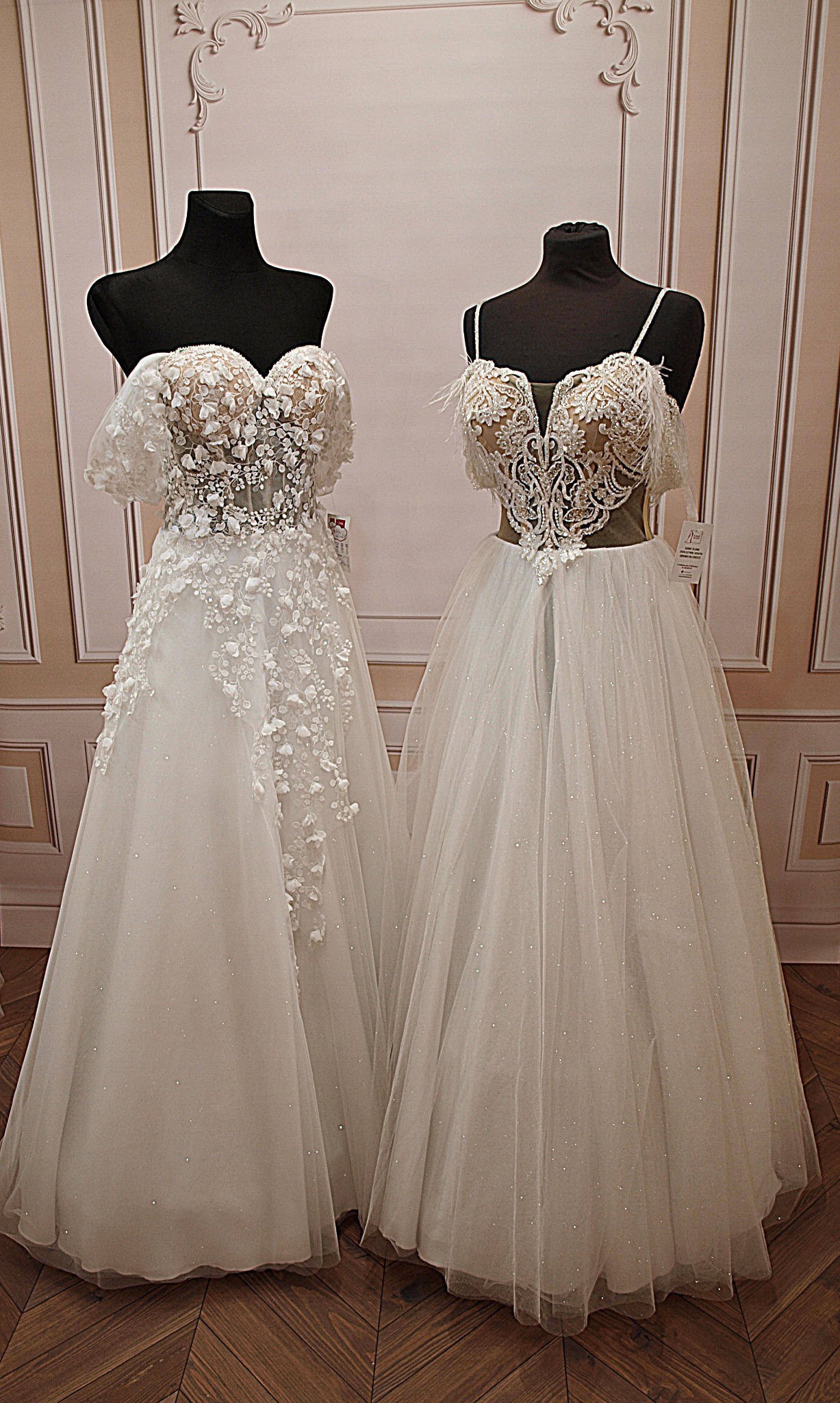 Dwie suknie ślubne na manekinach z czarnymi torsami. Suknie z koronkowymi zdobieniami i tiulowymi spódnicami, w tle ściana z dekoracyjnymi listwami i drewniana podłoga.