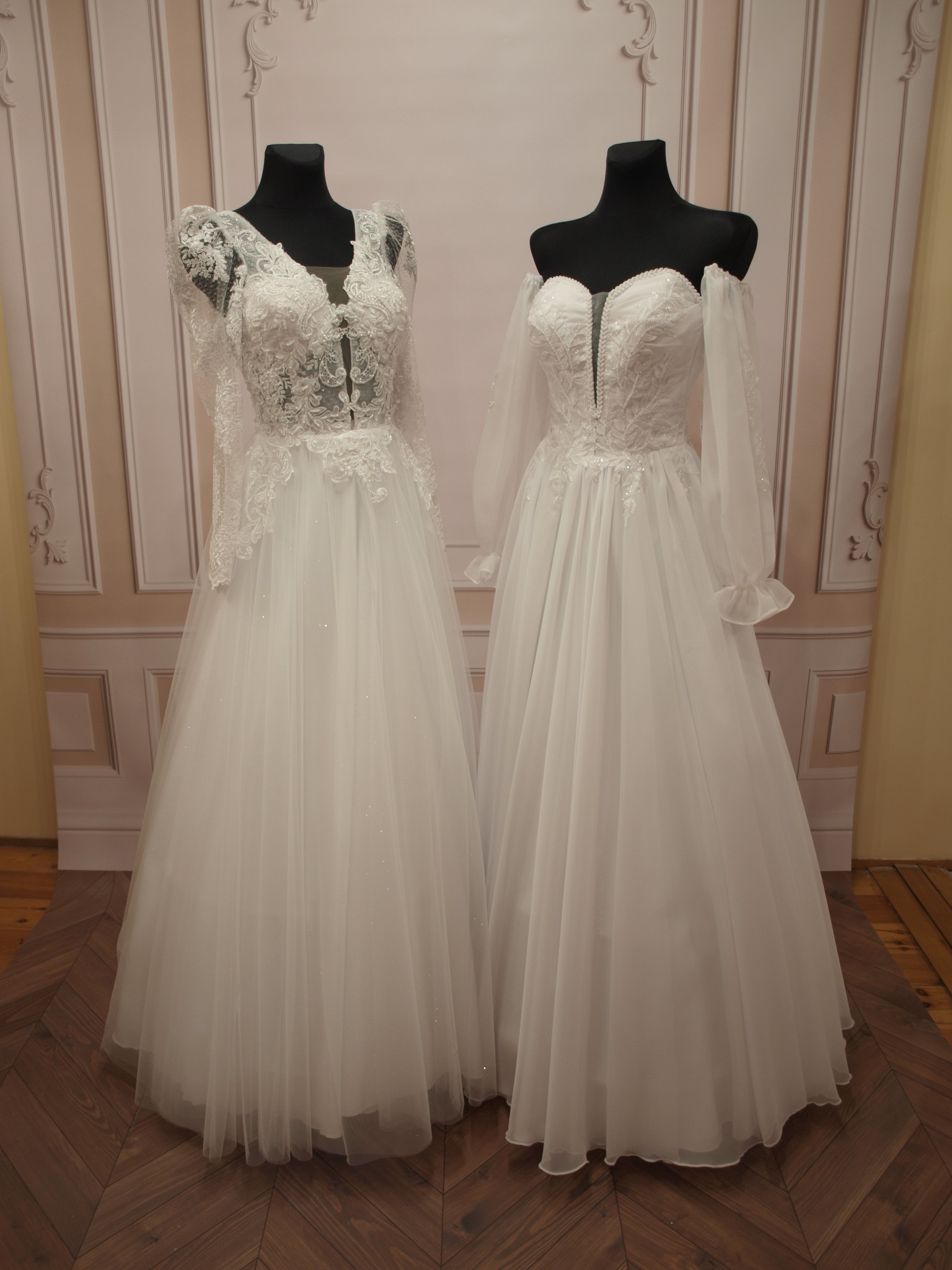 Dwie białe suknie ślubne z koronkowymi detalami na manekinach, na tle ściany w pastelowe wzory, prezentowane na drewnianej podłodze w jodełkę.