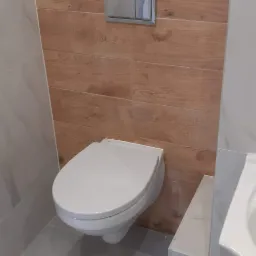 Nowoczesna, podwieszana toaleta z drewnopodobną zabudową i chromowanym przyciskiem spłukującym, na tle marmurowych płytek i szarej podłogi.