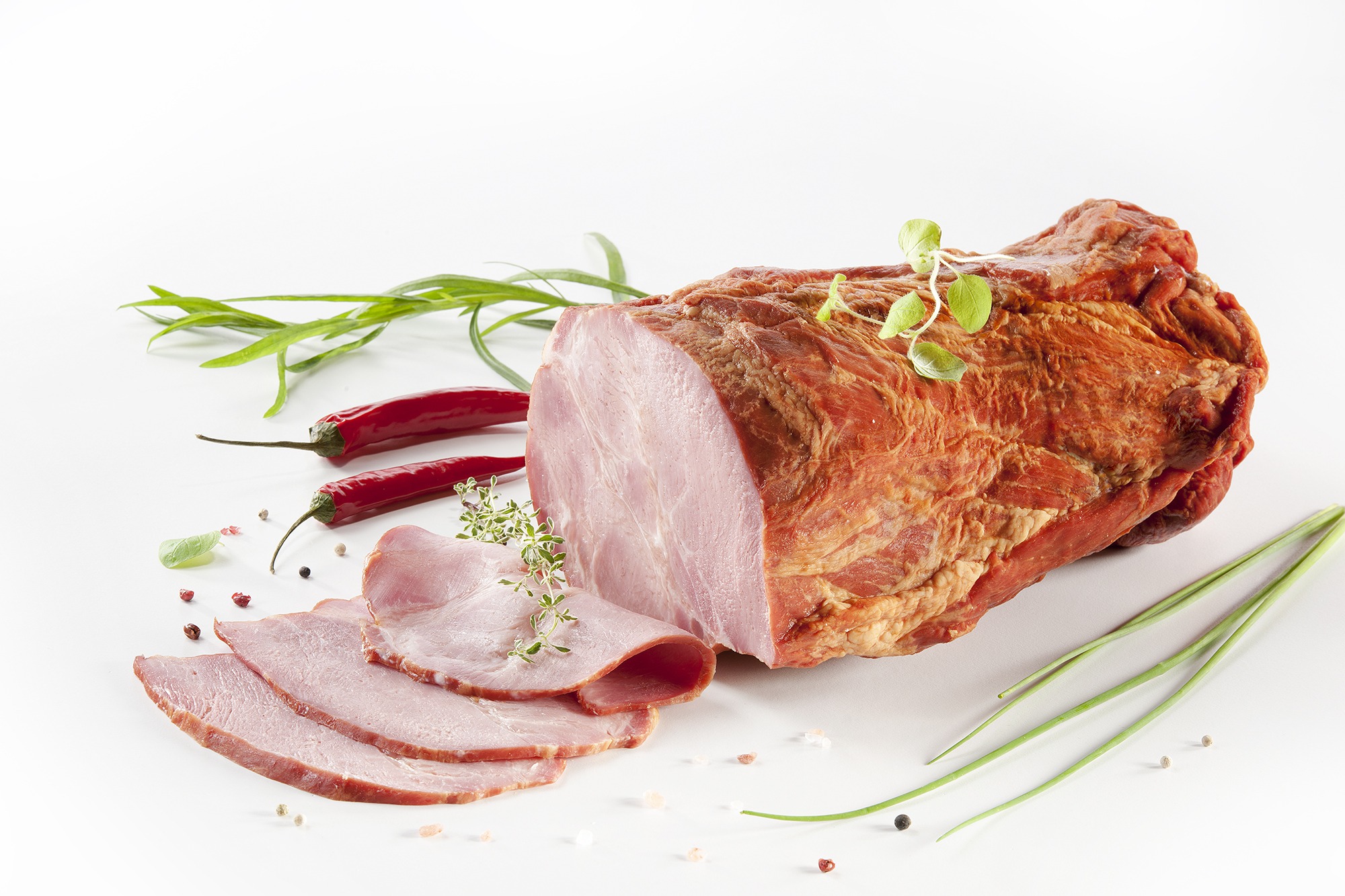 Kawałek wędzonego baleronu z plasterkami, ozdobiony gałązkami ziół, papryczkami chili i szczypiorkiem na białym tle.
