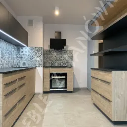 Nowoczesna kuchnia z drewnianymi frontami szafek, czarnymi blatami, wzorzystą mozaiką nad blatem i otwartymi półkami.