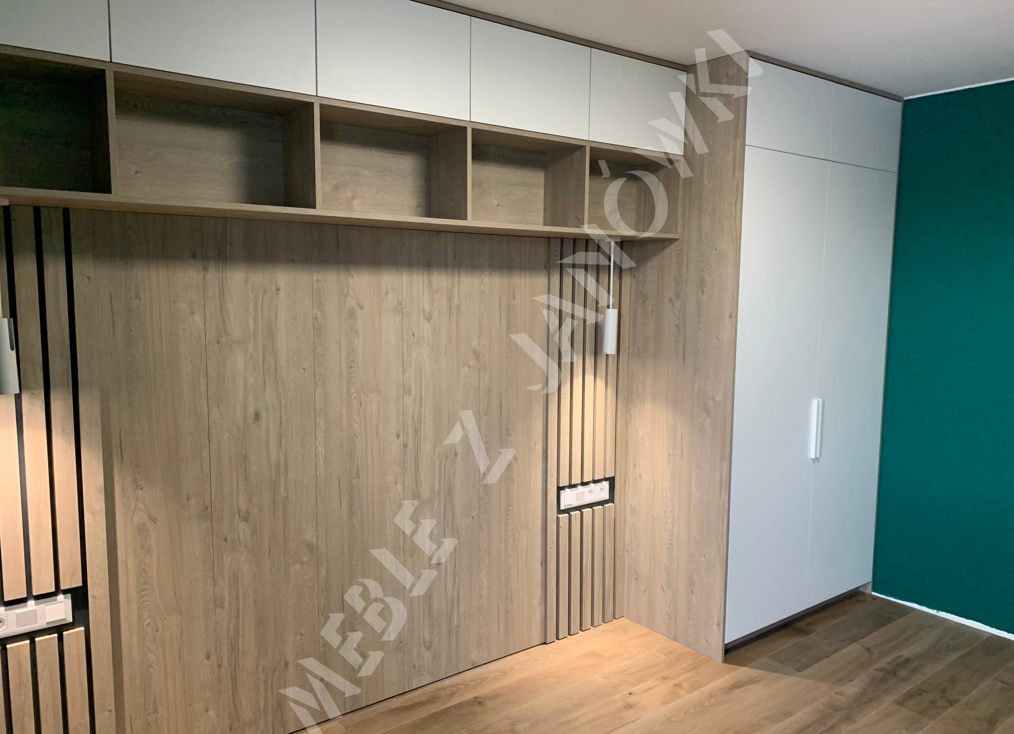 Nowoczesna zabudowa pokoju z drewnianą okładziną, białymi szafkami górnymi i wnękami na książki, uzupełniona białą szafą i zieloną ścianą w tle.