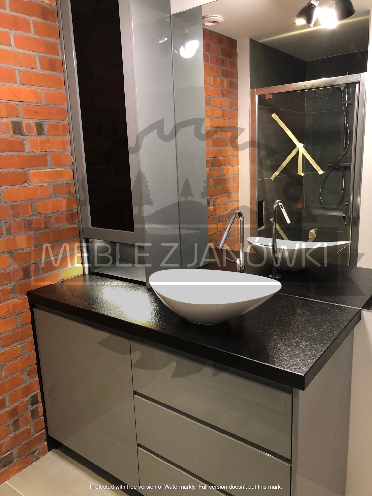 Łazienka w stylu loftowym z umywalką nablatową, szafką pod umywalkę z szarymi frontami i blatem z ciemnego kamienia, widoczna ściana z czerwonej cegły i lustro odbijające kabinę prysznicową.