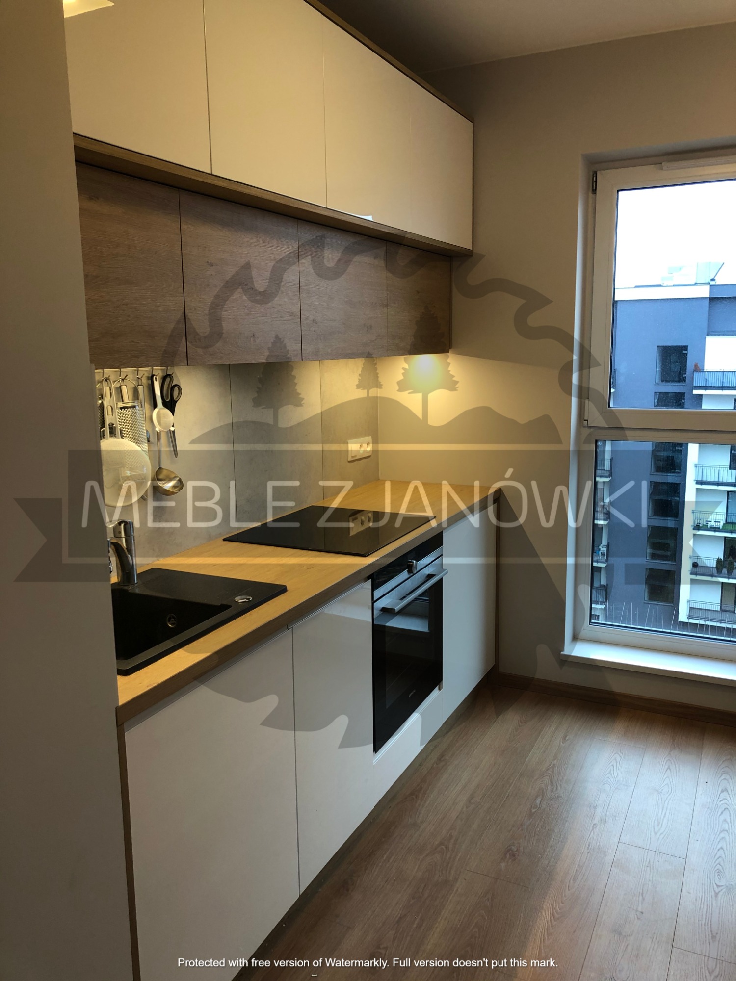 Nowoczesna kuchnia z białymi szafkami dolnymi i drewnianymi górnymi, czarny zlew i płyta indukcyjna na drewnianym blacie, widok z okna na budynek mieszkalny.