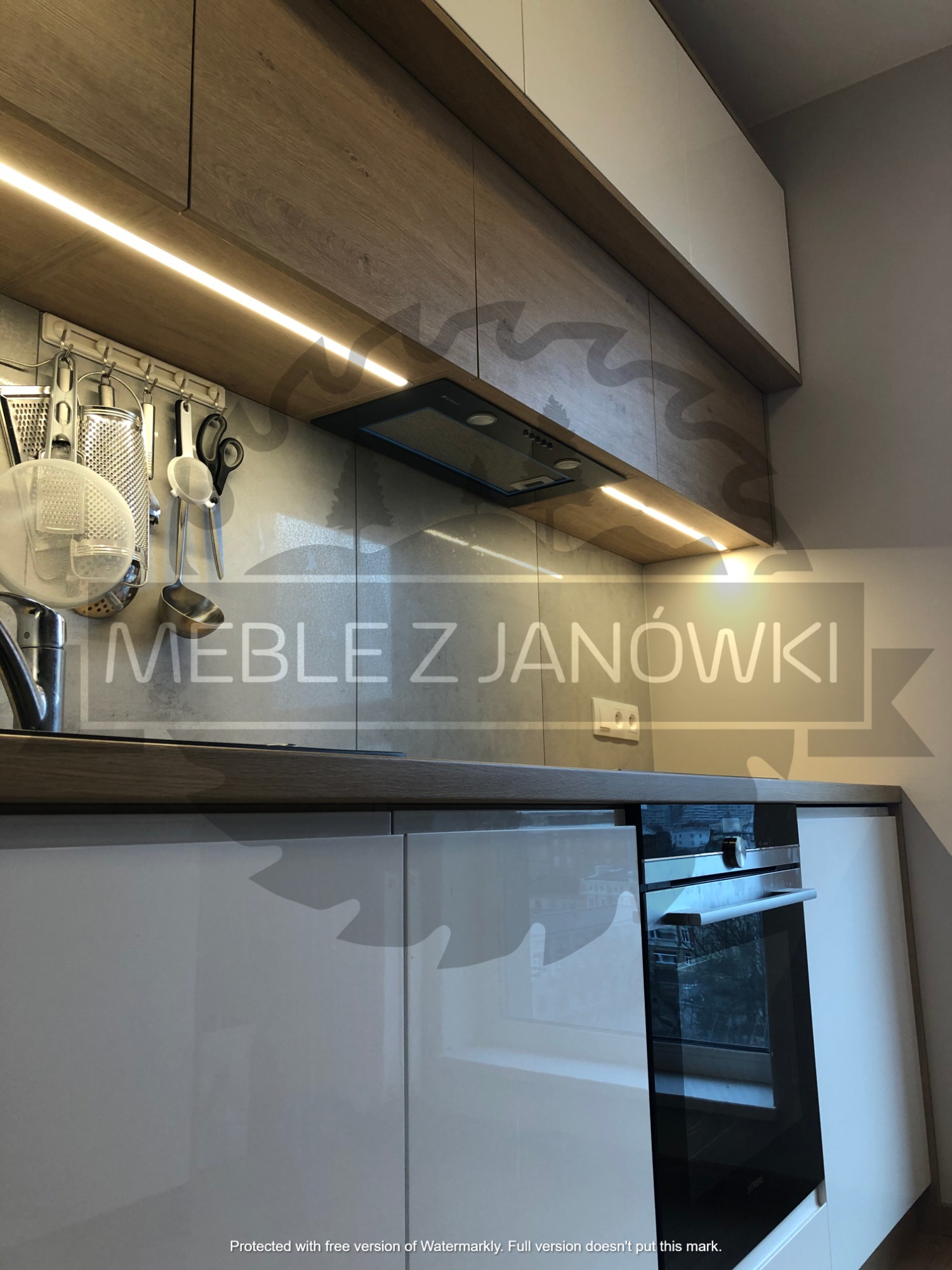 Nowoczesna kuchnia z białymi i drewnopodobnymi szafkami, podświetleniem LED podszafkowym, szarym blatem, relingiem kuchennym z akcesoriami i piekarnikiem w zabudowie.