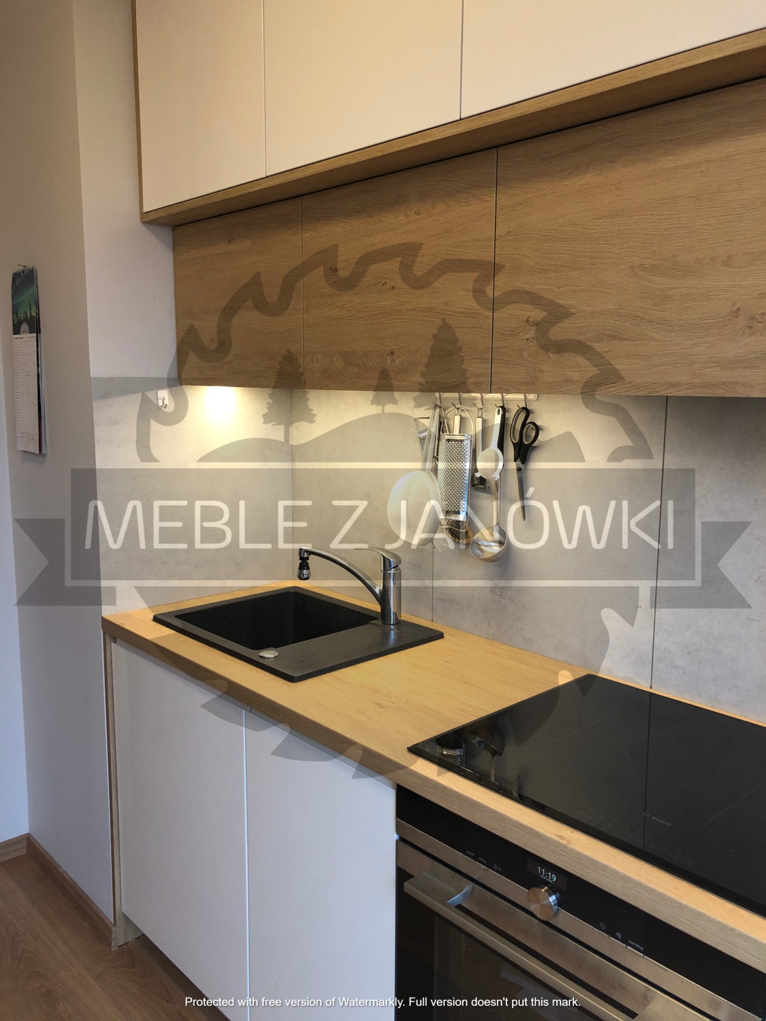 Nowoczesna kuchnia z drewnianym blatem, czarnym zlewem i płytą indukcyjną; białe szafki dolne i górne z drewnianymi frontami, reling z akcesoriami kuchennymi na szarej ścianie.