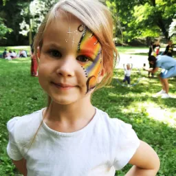 Uśmiechnięta dziewczynka z pomalowaną twarzą w motywy motyla i gwiazdki, na tle zielonego trawnika i osób w tle.