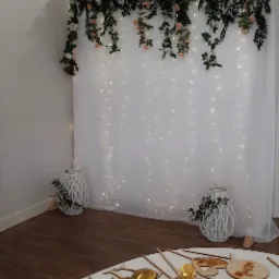 Dekoracja ślubna z białym tłem z delikatnymi światełkami, girlandą z kwiat&oacute;w i liści, białymi lampionami oraz złotymi balonami w kształcie cyfr i drewnianymi rekwizytami do zdjęć.