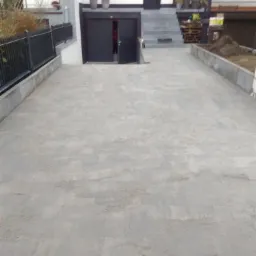 Szara, prostokątna nawierzchnia przed domem z garażem, wykonana z drobnych elementów, z widocznymi śladami piasku spomiędzy elementów, otoczona betonowymi obrzeżami.