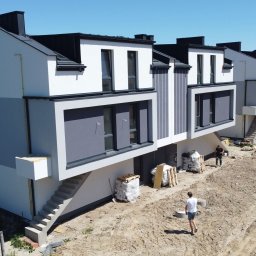 PHU Papierowski - Nowoczesne, szeregowe domy z szarą elewacją i czarnym dachem, widoczne z góry. Na pierwszym planie teren budowy z materiałami i dwoma osobami.