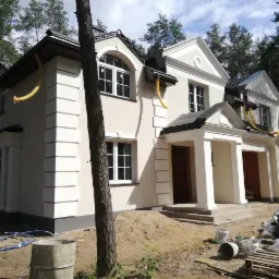 Nowo wykończony dom z kremową elewacją i białymi detalami architektonicznymi, widoczne żółte rury wentylacyjne wystające z okapów i nieporządek na placu budowy.