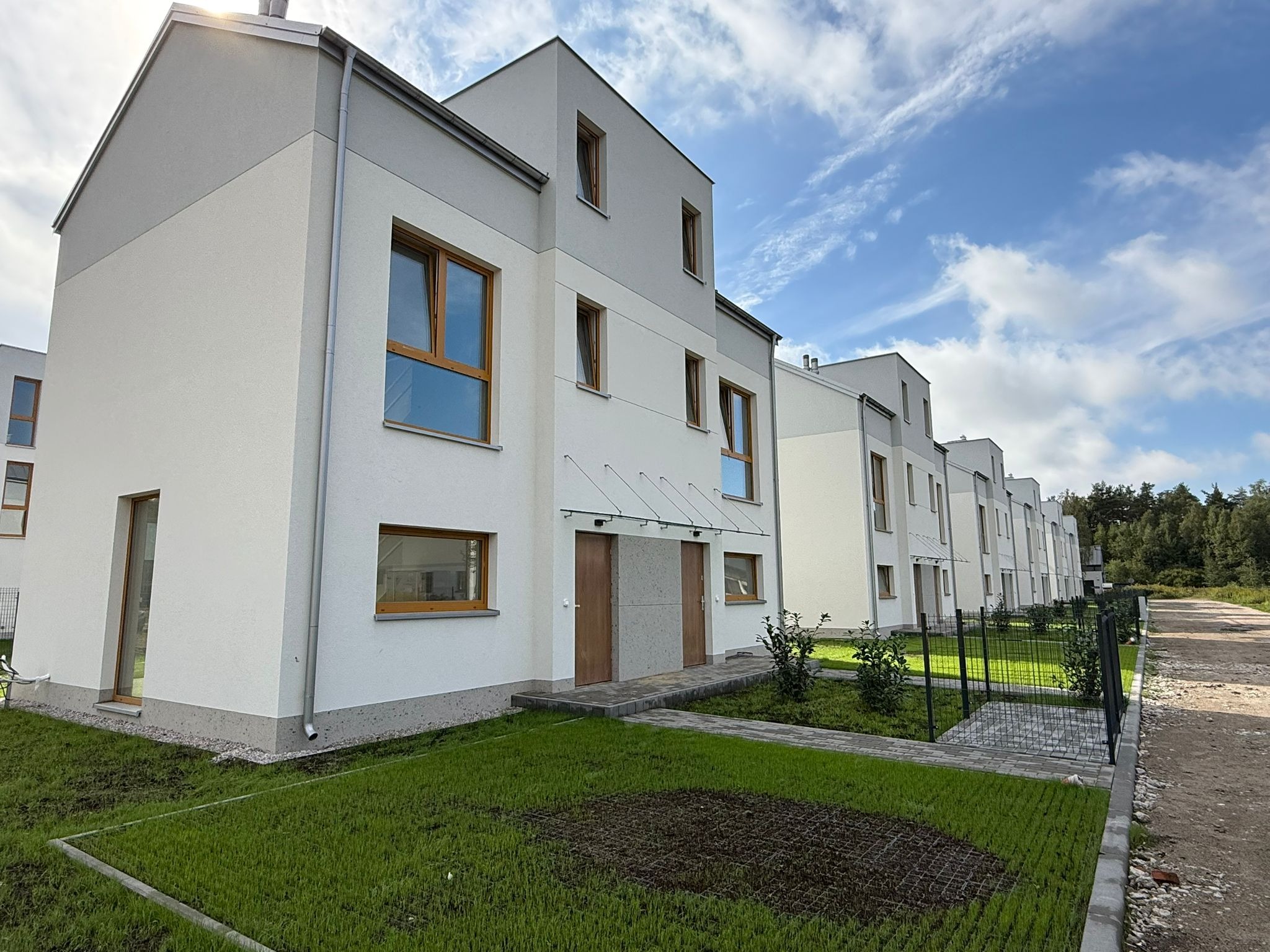 Szereg nowych, dwukondygnacyjnych domów z jasną elewacją i drewnianymi oknami, widocznych z perspektywy trawnika. Błękitne niebo z chmurami w tle.