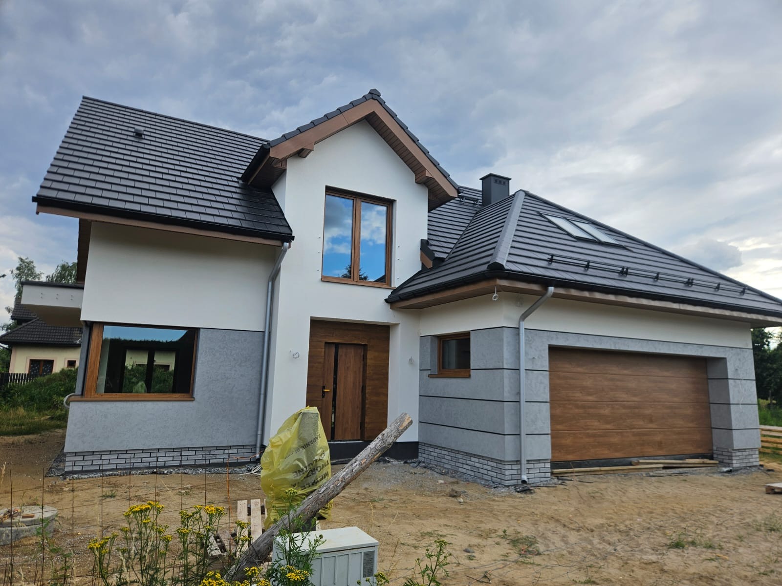 Nowoczesny dom jednorodzinny z białą elewacją i szarymi akcentami, brązowe okna i brama garażowa, dachówka w kolorze grafitu. Widok z poziomu gruntu.