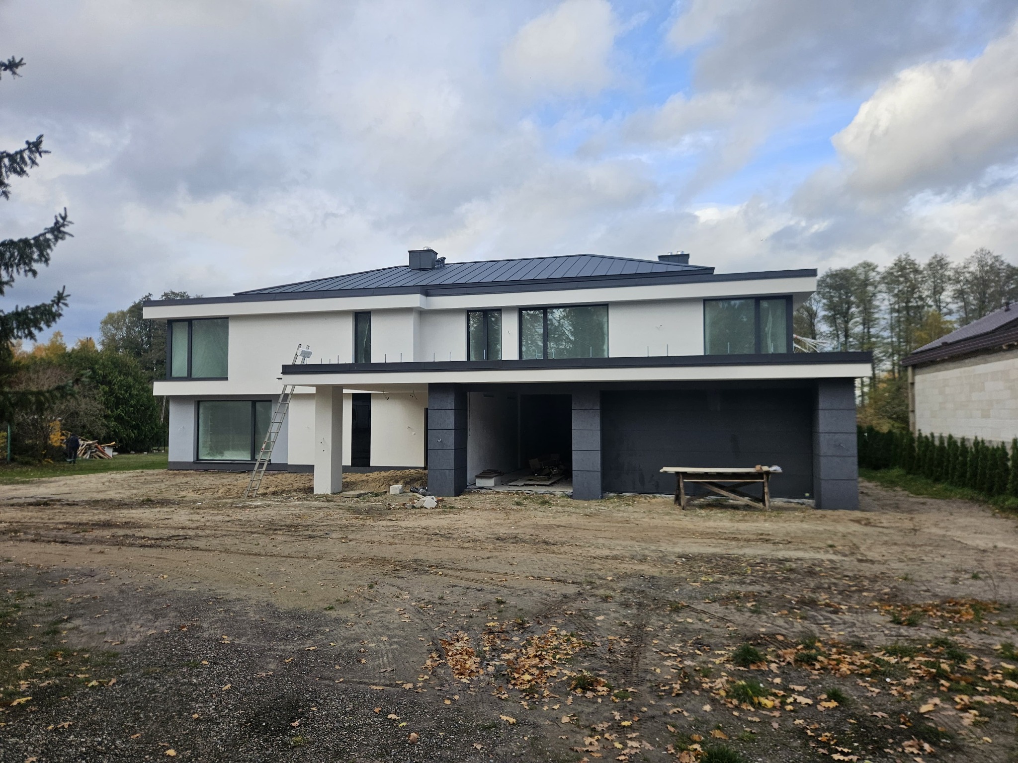 Nowoczesny, dwukondygnacyjny dom z płaskim dachem w trakcie wykończenia elewacji. Widoczna drabina oparta o ścianę, surowy teren wokół budynku.