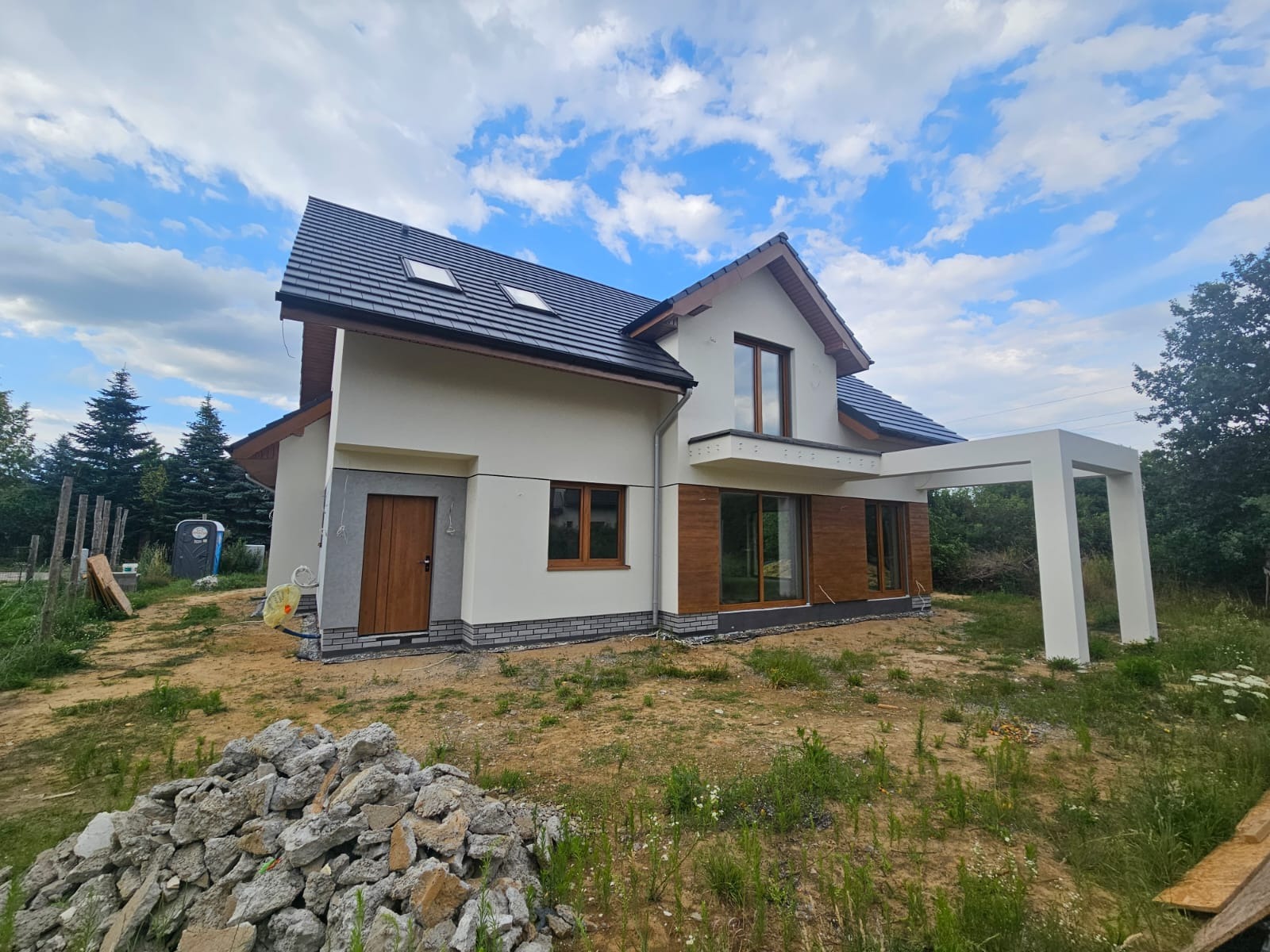 Nowoczesny dom jednorodzinny z jasną elewacją, drewnianymi akcentami i ciemnym dachem na tle błękitnego nieba z chmurami. Widoczny fragment ogrodu i sterta gruzu.