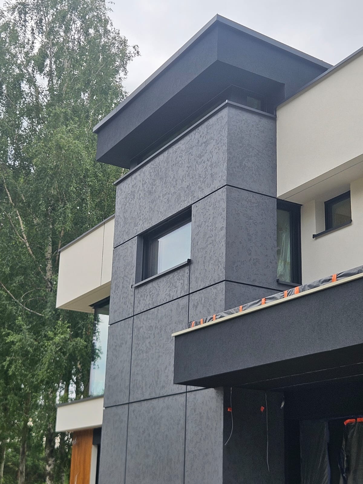 Nowoczesny dom z elewacją w odcieniach szarości i bieli, z widoczną fakturą tynku i elementami architektonicznymi. Ujęcie z dołu, częściowo widoczne drzewa w tle.