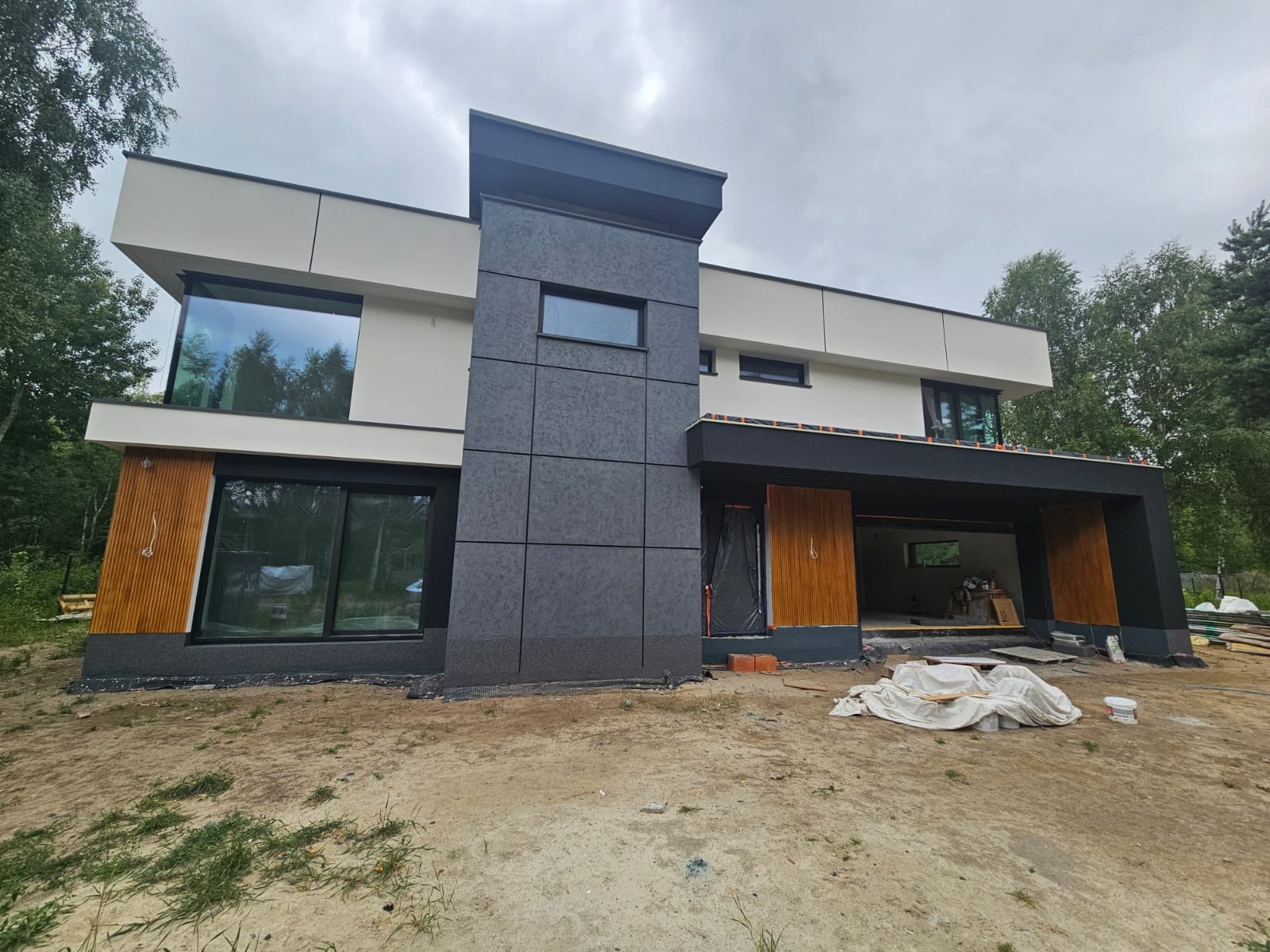 Nowoczesny dom z elewacją w odcieniach szarości i beżu, z elementami drewnianymi. Widoczne duże okna i częściowo wykończony podjazd. Dom otoczony zielenią.