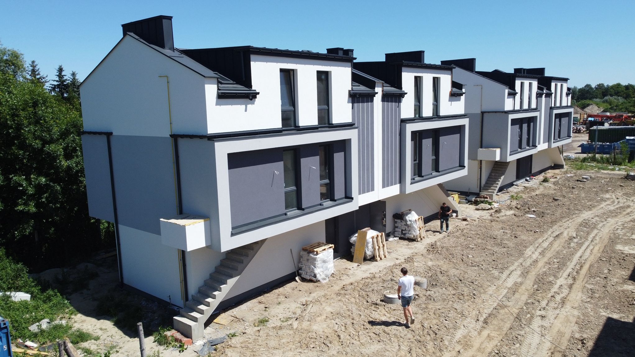 Nowoczesne, szeregowe domy z szarą elewacją i czarnym dachem, widoczne z góry. Na pierwszym planie teren budowy z materiałami i dwoma osobami.
