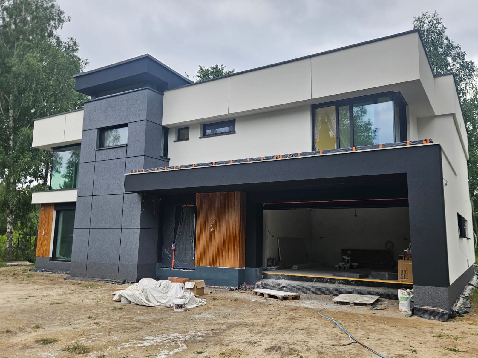 Nowoczesny dom w Starych Babicach z elewacją w odcieniach szarości i bieli, częściowo wykończony, z widocznymi materiałami budowlanymi na posesji.