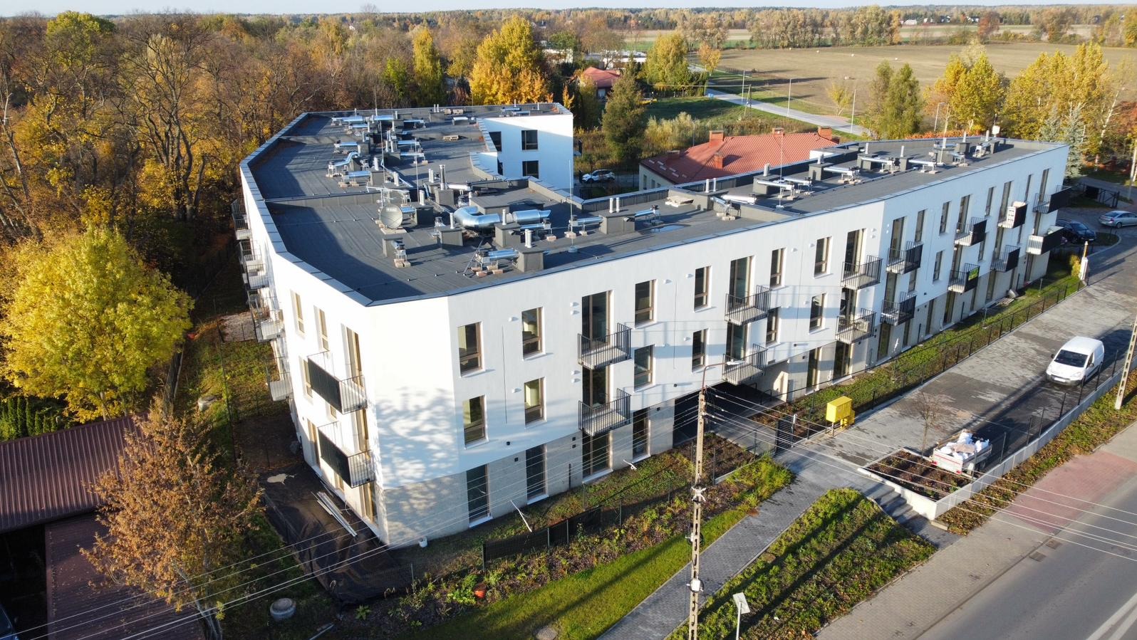 Nowoczesny, biały budynek wielorodzinny z balkonami, widziany z lotu ptaka w jesiennej scenerii. Charakterystyczne elementy wentylacyjne na dachu.