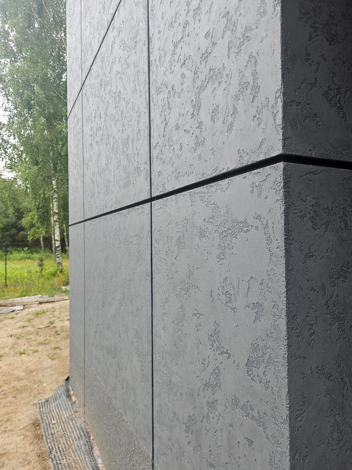 Szara elewacja budynku z fakturą, wykonana z paneli. Widoczny narożnik budynku i fragment zieleni w tle. Elewacja o nowoczesnym wyglądzie.