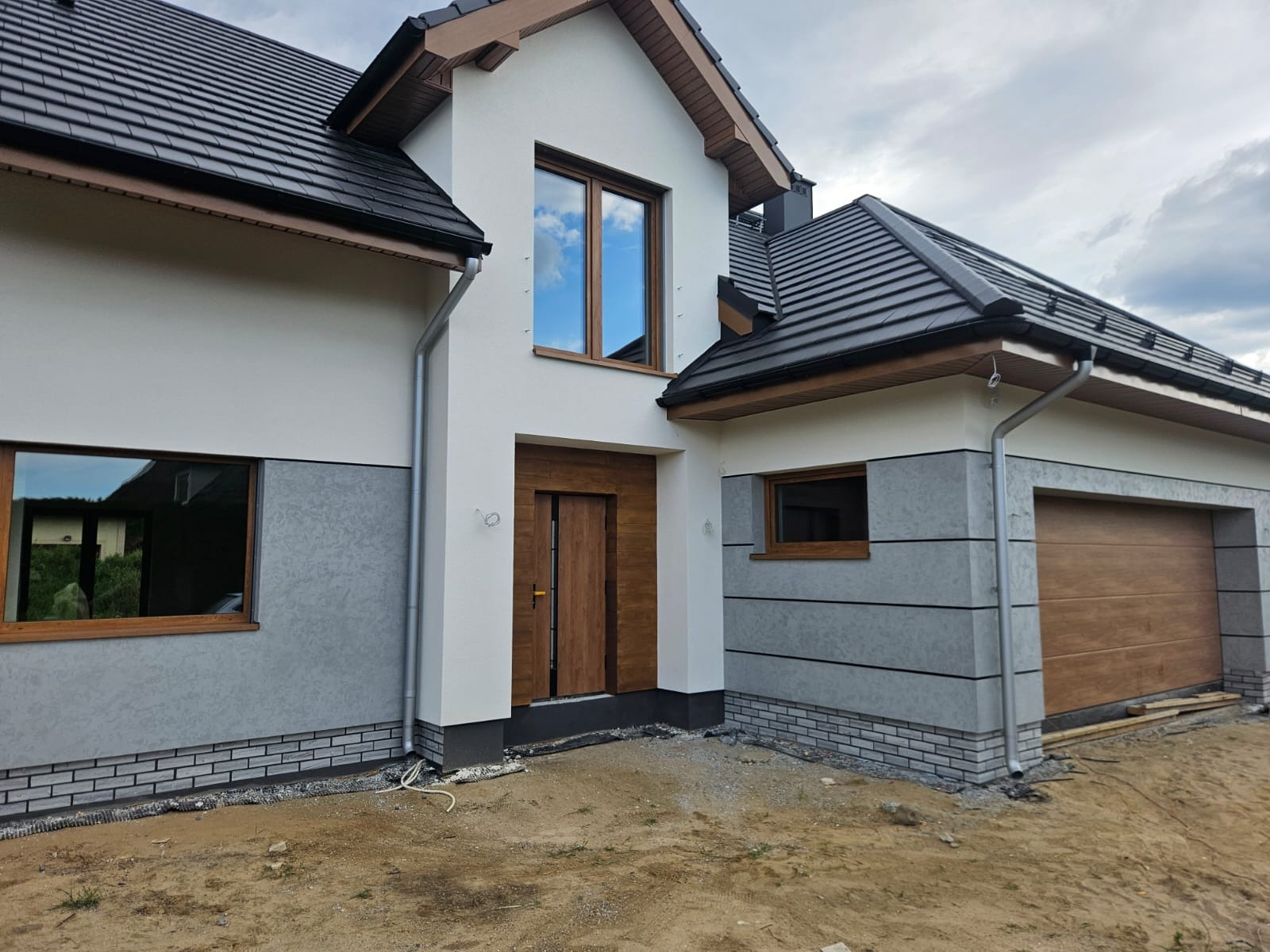 Nowoczesny dom jednorodzinny z jasną elewacją, grafitowym dachem i drewnianymi oknami. Widoczny fragment garażu z bramą segmentową w kolorze drewna.
