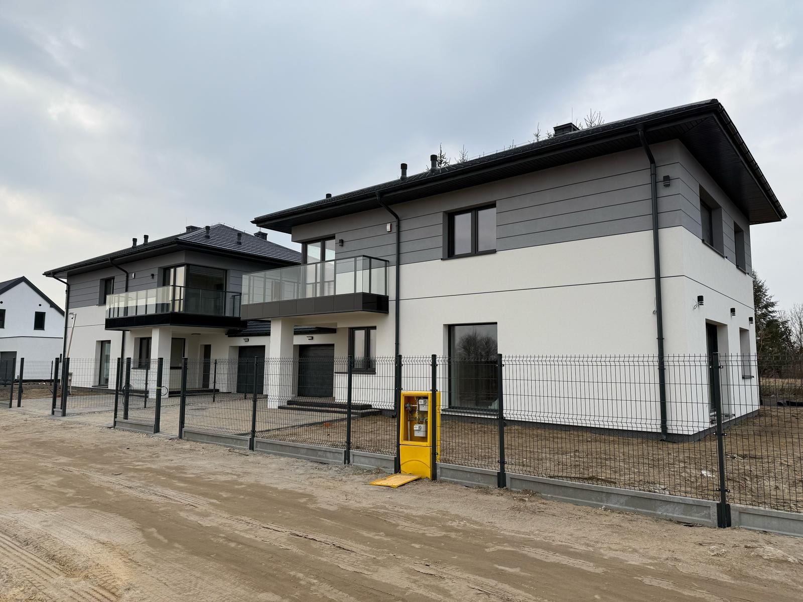 Dwa nowoczesne, dwukondygnacyjne domy z balkonami, wykończone w bieli i szarości, ogrodzone siatką. Na pierwszym planie żółta skrzynka gazowa.