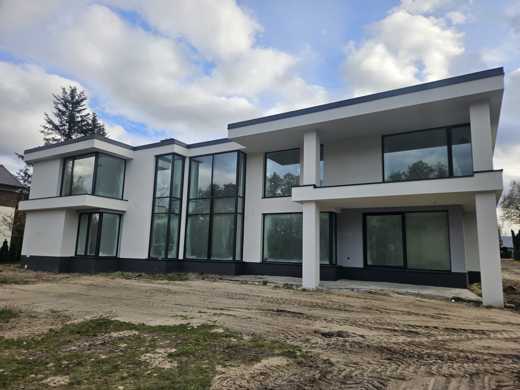 Nowoczesny, dwukondygnacyjny dom z białymi ścianami i dużymi oknami z czarnymi ramami, widoczny z perspektywy ziemi, na tle błękitnego nieba z chmurami.